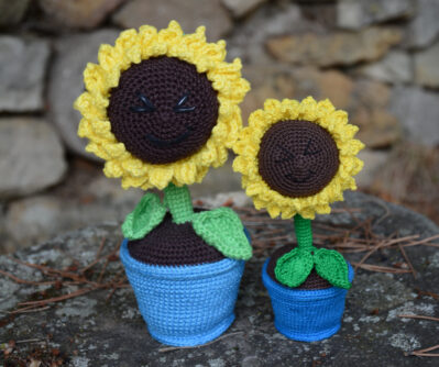 Deux tournesols crochetés de tailles différentes, placés côte à côte sur un rocher. Chaque fleur est dans un pot bleu, avec une tige verte et un cœur brun entouré de pétales jaunes. Les deux tournesols affichent un visage souriant brodé avec des yeux rieurs fermés. À l'arrière-plan, un mur de pierres.