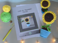 Des pages du patron de lulu le tournesol sont posés sur un fond gris. Sur le côté droit de ces pages se trouve 2 versions crochetées de Lulu le tournesol, une plus grande couchée et l'autre plus petite. L'un des tournesol est posé couché dans son pot, l'autre est debout dans son pot. Les 2 ont le visage orientés vers le haut, semblant nous sourire. À l'avant-plan se trouve une guirlande lumineuse.