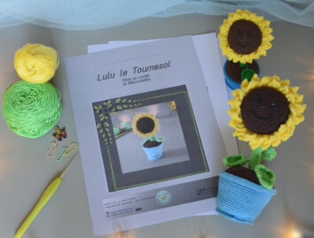Des pages du patron de lulu le tournesol sont posés sur un fond gris. Sur le côté droit de ces pages se trouve 2 versions crochetées de Lulu le tournesol, une plus grande couchée et l'autre plus petite. L'un des tournesol est posé couché dans son pot, l'autre est debout dans son pot. Les 2 ont le visage orientés vers le haut, semblant nous sourire. À l'avant-plan se trouve une guirlande lumineuse.
