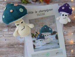 Des pages du patron d'Ottie le champignon sont posées sur une table en bois. Ces pages sont entourées d'un côté et de l'autre de deux versions du champignon, une grande sur la gauche et une plus petite sur la droite. Le grand champignon a un chapeau bleu pétrole et des pois spots rose et vert d'eau. Le petit lui a un chapeau violet décoré de spots coloré. En fond il y a quelques feuilles automnales ainsi qu'un marron. Une guirlande lumineuse entoure le tout.