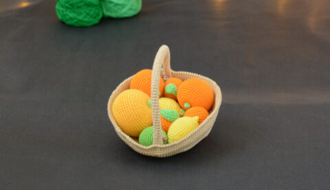 sur un fond gris foncé, il y a un panier crocheté en topaze garni d'agrumes : citrons, citron vert, clémentines, oranges et pamplemousse. À l'arrière plan, sur la gauche il y a 2 pelotes vertes. En fond est disposé un nuage de ouate et une guirlande lumineuse.