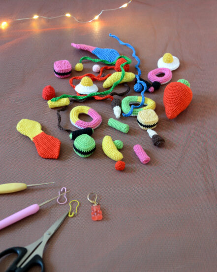 Un lot de bonbons crochetés disposés sur un fond orangé. On y trouve des bananes, des œufs au plat, des frites, des réglisses, un grosse fraise, et d'autres encore. À l'arrière-plan, dans le coin supérieur gauche se trouve une guirlande lumineuse. À l'avant-plan, dans le coin inférieur droit sont disposés des crochets, ciseaux et anneaux marqueurs, ils pointent en direction des bonbons.