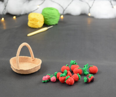 sur un fond gris foncé, sur la gauche il y a un panier crocheté en topaze, et sur sa droite se trouvent des fruits rouges miniatures : fraises, cerises et baies variées (framboises, mûres,...). À l'arrière plan, sur la gauche il y a 2 pelotes jaune et verte et un crochet. En fond est disposé un nuage de ouate et une guirlande lumineuse.
