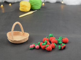 Panier de fruits rouges 12 lum-16 sur un fond gris foncé, sur la gauche il y a un panier crocheté en topaze, et sur sa droite se trouvent des fruits rouges miniatures : fraises, cerises et baies variées (framboises, mûres,...). À l'arrière plan, sur la gauche il y a 2 pelotes jaune et verte et un crochet. En fond est disposé un nuage de ouate et une guirlande lumineuse.