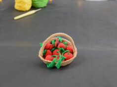 sur un fond gris foncé, il y a un panier crocheté en topaze garni de fruits rouges miniatures : fraises, cerises et baies variées (framboises, mûres,...). À l'arrière plan, sur la gauche il y a 2 pelotes jaune et verte et un crochet. En fond est disposé un nuage de ouate et une guirlande lumineuse.