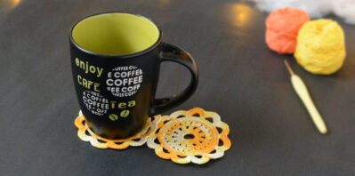 Une tasse noire à l'intérieur vert clair est posée sur deux sous-verres ronds crochetés dans un dégradé jaune et orange. En fond, à droite, deux pelotes de coton (jaune et orange) et un crochet. En arrière-plan, des guirlandes lumineuses sont disposées sur du rembourrage blanc façon nuages.