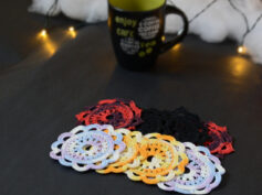Huit sous-verres ronds crochetés sont posés sur un fond gris sombre. Ils sont de différentes couleurs (dégradé parme et lilas, dégradé orange et jaune, dégradé violet et aubergine et uni noir. En fond il y a une tasse noire à l'intérieur vert clair et des guirlandes lumineuses sont disposées sur du rembourrage blanc façon nuages.