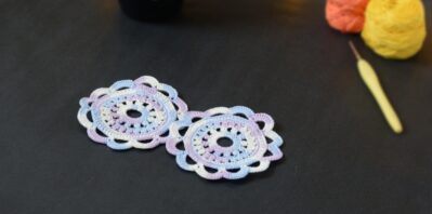 Deux sous-verres ronds crochetés dans un dégradé parme et lilas sont posés sur un fond gris sombre. En fond, à droite, deux pelotes de coton (jaune et orange) et un crochet. Sur la gauche, il y a une tasse noire à l'intérieur vert clair. En arrière-plan, des guirlandes lumineuses sont disposées sur du rembourrage blanc façon nuages.
