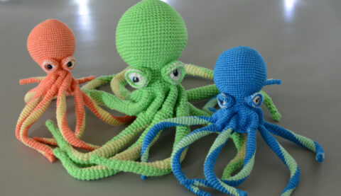 trio de poulpe crocheté les uns à côté des autres avec leurs tentacules entrelacés. Le plus à gauche est corail avec des tentacules corail et jaune pastel, celui du milieu est plus grand que les 2 autres, il est vert clair avec des tentacules vert clair et vert anis. le plus à droite est bleu, aux tentacules bleues et vert d'eau. Ils sont posés sur un fond gris, en fond il y a une guirlande lunimeuse ainsi qu'un nuage de ouate.