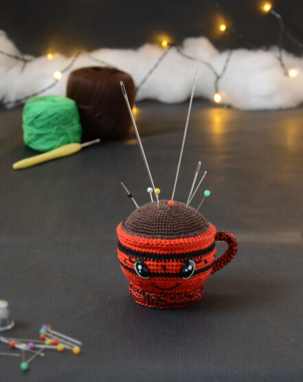 Porte-épingles crocheté en forme de tasse rouge. Le haut de la tasse est marron pour représenter une boisson chaude. La tasse est piquée d’aiguilles et d’épingles colorées. Elle a des yeux brillants et un sourire et est dans un décor gris foncé illuminé par une guirlande et des nuages de ouate. En arrière-plan, deux pelotes verte et marron et un crochet. À l'avant-plan, sur la gauche, il y a des épingles et un dé à coudre.
